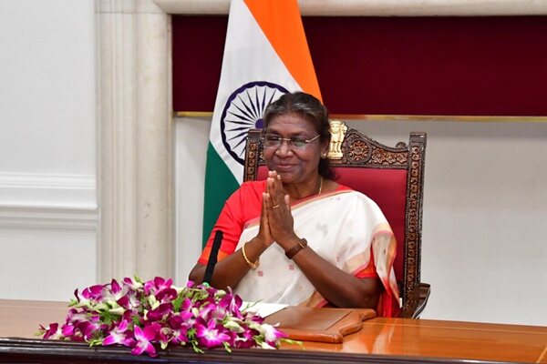 President Droupadi Murmu seated at Rashtrapati Bhavan with hands folded in greeting, the national flag behind her