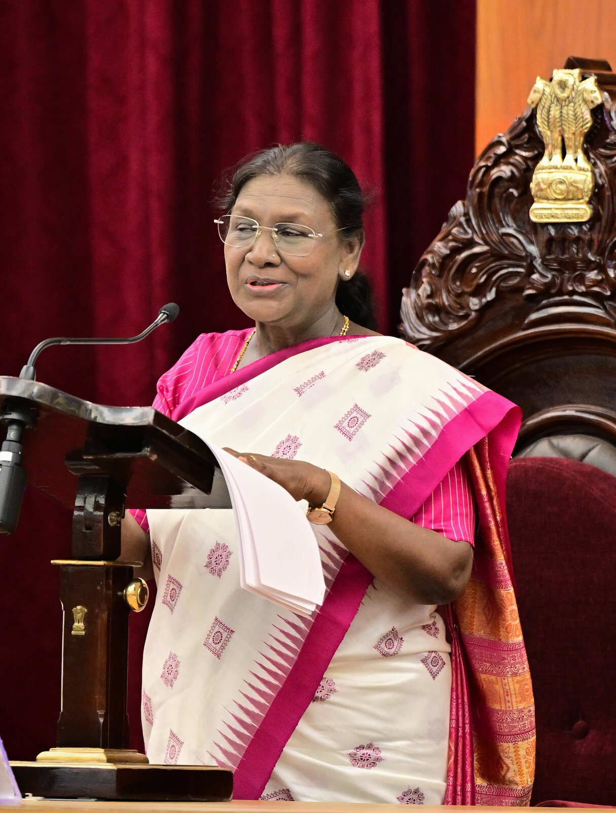 President Droupadi Murmu delivering an address from a wooden lectern, beside a ceremonial chair bearing the State Emblem of India