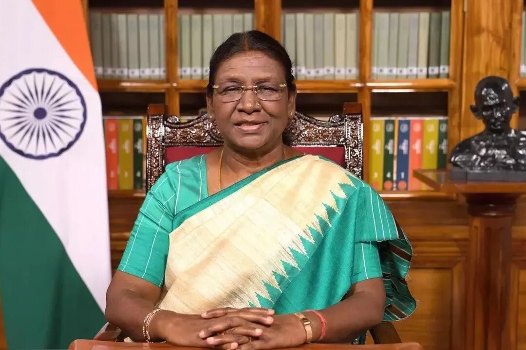 President Droupadi Murmu delivering an address to the nation from the library at Rashtrapati Bhavan, with the national flag and a bust of Mahatma Gandhi